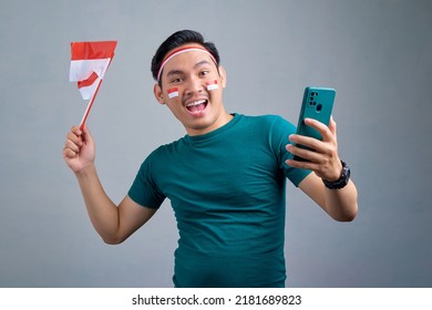 Excited young Asian man holding mobile phone and indonesian flag while celebrating indonesia independence day isolated on grey background. indonesia independence day celebration concept