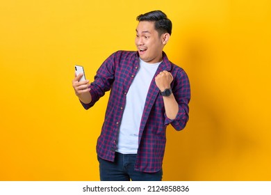 Excited Young Asian Man In Plaid Shirt Using Smartphone And Doing Winner Gesture Isolated On Yellow Background