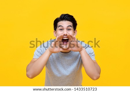 Excited young Asian man shouting with hands cupped around mouth isolated on yellow background