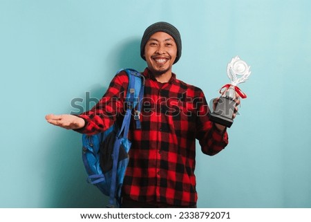Excited Young Asian man student wearing a backpack, beanie hat, and red plaid flannel shirt, holding trophy, smiling at the camera, isolated on a blue background