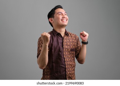 Excited Young Asian Man Wearing Batik Shirt Celebrating Victory And Raised Fists Isolated On Grey Background