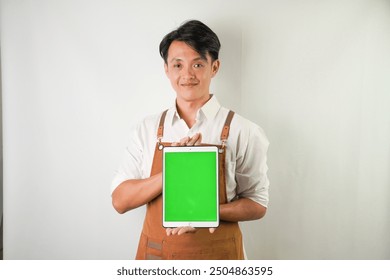 Excited young asian man wearing rolled-up white sleeve shirt and brown apron is showing vertical blank green screen tablet with smiling face expression. Isolated over white background. - Powered by Shutterstock