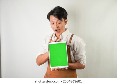 Excited young asian man wearing rolled-up white sleeve shirt and brown apron is showing vertical blank green screen tablet with smiling face expression. Isolated over white background. - Powered by Shutterstock