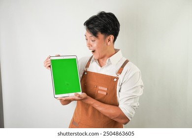 Excited young asian man wearing rolled-up white sleeve shirt and brown apron is showing vertical blank green screen tablet with smiling face expression. Isolated over white background. - Powered by Shutterstock