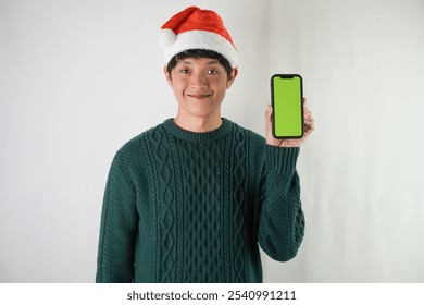 Excited young asian man wearing santa clause hat and green long sleeved sweater is showing smartphone green screen, isolated over white background. Concept for Christmas Holiday and New Year Party - Powered by Shutterstock