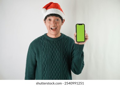 Excited young asian man wearing santa clause hat and green long sleeved sweater is showing smartphone green screen, isolated over white background. Concept for Christmas Holiday and New Year Party - Powered by Shutterstock