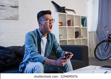 Excited Young Asian Man Yelling And Playing Video Game With Joystick At Home 