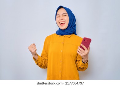 Excited Young Asian Muslim Woman Dressed In Orange Shirt Using Mobile Phone, Celebrating Success, Getting Good News Isolated Over White Background