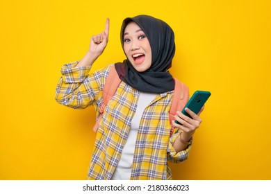 Excited Young Asian Muslim Woman Student In Plaid Shirt And Backpack Holding Mobile Phone, Pointing Finger Up, Creating Genius Solutions Isolated On Yellow Background