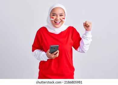 Excited young Asian muslim woman in red white t-shirt holding mobile phone and celebrating indonesian independence day on 17 august isolated on white background
