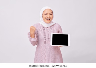 Excited young Asian Muslim woman wearing hijab and purple dress  showing digital tablet with empty screen for mockup and celebrating success isolated on white studio background - Powered by Shutterstock