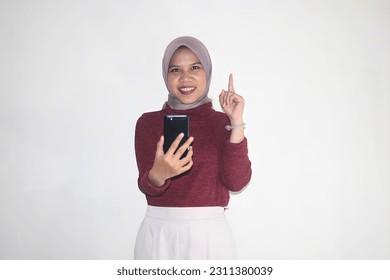 Excited young Asian Muslim woman with hijab holding mobile phone and pointing finger up, creating genius solutions isolated over white background. - Shutterstock ID 2311380039