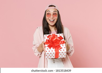 Excited young asian woman in casual clothes cap glasses isolated on pastel pink wall background. St. Valentine's Day, Women's Day, birthday, holiday concept. Hold red present box with gift ribbon bow
