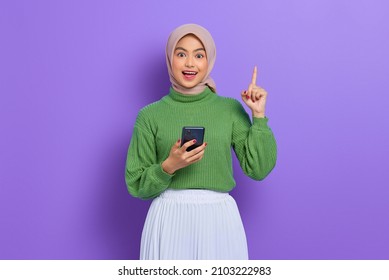 Excited Young Asian Woman In Green Sweater And Hijab Holding Mobile Phone And Pointing Finger Up, Creating Genius Solutions Isolated Over Purple Background