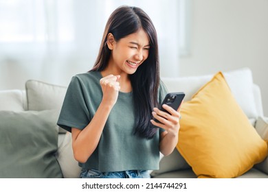 Excited young asian woman hold phone chatting in dating app feel happy sit on sofa at home, Closeup joyful female reading good news on phone. Surprised lady celebrating victory on phone - Shutterstock ID 2144744291
