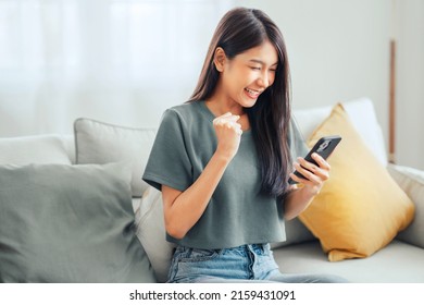 Excited young asian woman hold phone chatting in dating app feel happy sit on sofa at home, Closeup joyful female reading good news on phone. Surprised lady celebrating victory on phone - Shutterstock ID 2159431091