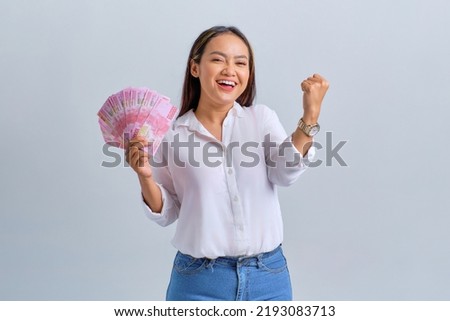 Excited young Asian woman holding money banknotes and making success gestures isolated over white background