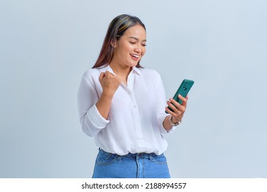 Excited Young Asian Woman Looking At Mobile Phone Screen Gesturing Yes With Raised Fist Reacting To Online News Isolated Over White Background