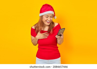 Excited Young Asian Woman In Santa Claus Hat Holding Cell Phone With Hands On Chest On Yellow Background