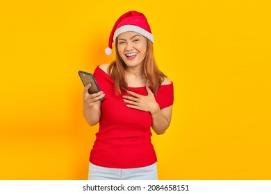 Excited Young Asian Woman In Santa Claus Hat Holding Cell Phone With Hands On Chest On Yellow Background