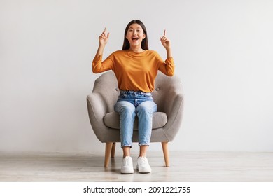 Excited young Asian woman sitting in armchair, pointing two fingers up, offering space for your advertisement against white studio wall, full length. Best promotion, cool discount, huge sale concept