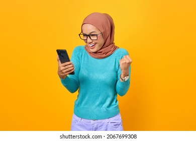 Excited young Asian woman using smartphone celebrating success isolated on yellow background