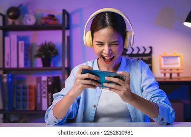 Excited Young Asian woman wearing headset and playing online game on smartphone with live broadcasting on internet.