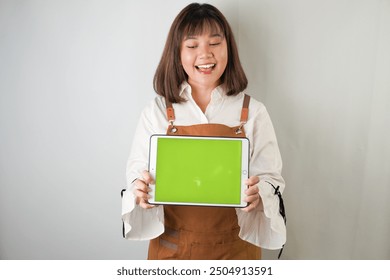 Excited young asian woman wearing white long sleeve shirt and brown apron is showing horizontal blank green screen tablet with smiling face expression. Isolated over white background. - Powered by Shutterstock