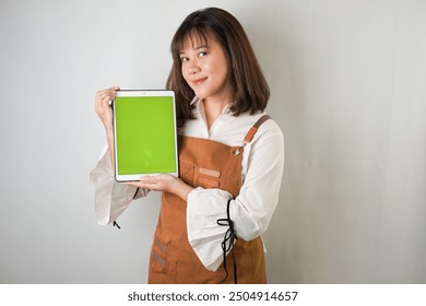 Excited young asian woman wearing white long sleeve shirt and brown apron is showing vertical blank green screen tablet with smiling face expression. Isolated over white background. - Powered by Shutterstock