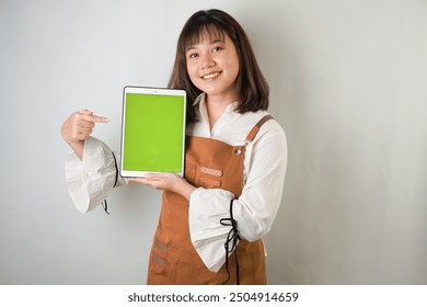 Excited young asian woman wearing white long sleeve shirt and brown apron is showing vertical blank green screen tablet with smiling face expression. Isolated over white background. - Powered by Shutterstock