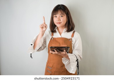 Excited Young asian woman wearing white long sleeve shirt and brown apron is holding a tablet and pointing upward with smiling face expression. Isolated over white background. - Powered by Shutterstock