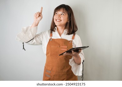 Excited Young asian woman wearing white long sleeve shirt and brown apron is holding a tablet and pointing upward with smiling face expression. Isolated over white background. - Powered by Shutterstock