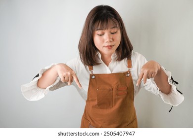 Excited young asian woman wearing long white sleeve shirt and brown apron is pointing downward with smiling face expression. Isolated over white background - Powered by Shutterstock