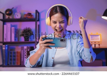 Excited Young Asian woman wearing headset and playing online game on smartphone at night modern house.