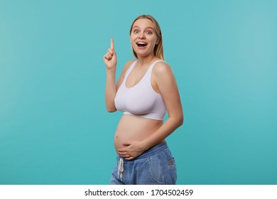 Excited Young Attractive White-headed Future Mom Looking At Camera With Wide Eyes And Mouth Opened While Standing Over Blue Background With Raised Forefinger