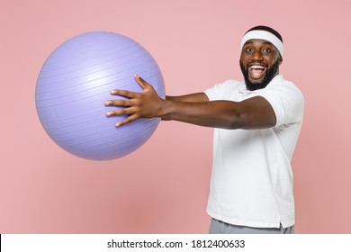 Excited Young Bearded African American Fitness Sports Man In White Headband T-shirt Posing Hold Fitball Looking Camera Spending Time In Gym Isolated On Pastel Pink Color Background Studio Portrait