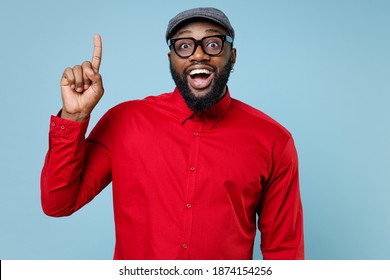 Excited Young Bearded African American Man In Casual Red Shirt Cap Eyeglasses Standing Holding Index Finger Up With Great New Idea Looking Camera Isolated On Pastel Blue Background Studio Portrait