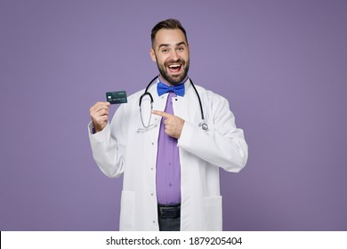 Excited Young Bearded Doctor Man In White Medical Gown Stethoscope Point Index Finger On Credit Bank Card Isolated On Violet Background Studio Portrait. Healthcare Personnel Health Medicine Concept