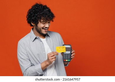 Excited young bearded Indian man 20s years old wears blue shirt hold wireless modern bank payment terminal to process acquire credit card payments isolated on plain orange background studio portrait
