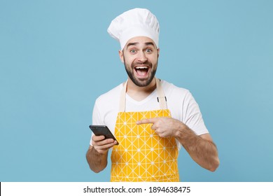 Excited Young Bearded Male Chef Or Cook Baker Man In Apron White T-shirt Toque Chefs Hat Isolated On Blue Wall Background Studio Portrait. Cooking Food Concept. Pointing Index Finger On Mobile Phone