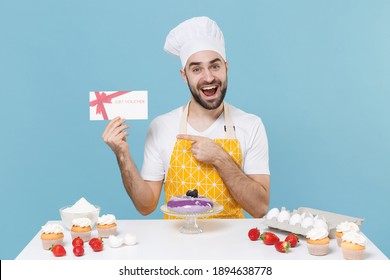 Excited young bearded male chef or cook baker man in apron white t-shirt toque chefs hat cooking at table isolated on blue background. Cooking food concept. Pointing index finger on gift certificate - Shutterstock ID 1894638778