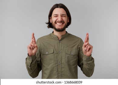 Excited young bearded male keeping his fingers crossed and closed his eyes tightly while making a wish. Positive man sincerely believing in his luck, hopes for the best, isolated on grey background