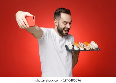 Excited young bearded man 20s in white t-shirt doing selfie shot on mobile phone hold makizushi sushi roll served on black plate traditional japanese food isolated on red background studio portrait