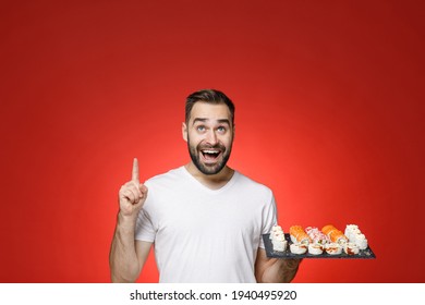 Excited Young Bearded Man In Basic White T-shirt Pointing Index Finger Overhead Up Hold Makizushi Sushi Roll Served On Black Plate Traditional Japanese Food Isolated On Red Background Studio Portrait