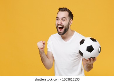 Excited young bearded man football fan in white t-shirt cheer up support favorite team with soccer ball screaming clenching fist isolated on yellow background. People sport family leisure concept.