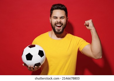 Excited young bearded man football fan in yellow t-shirt cheer up support favorite team hold in hand soccer ball doing winner gesture celebrate isolated on plain dark red background studio portrait
