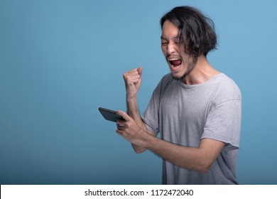 Excited Young Bearded Man Playing Mobile Games In Blue Background. Asian Young Hipster Playing Mobile Game And Win, Half Body Shot. Young Generation Hipster Relaxing Concept.