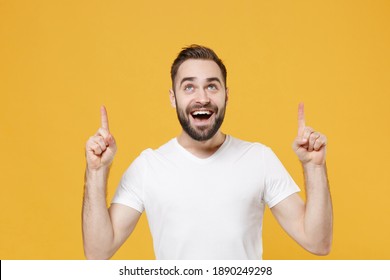 Excited Young Bearded Man In White Casual T-shirt Posing Isolated On Yellow Background Studio Portrait. People Sincere Emotions Lifestyle Concept. Mockup Copy Space. Pointing Index Fingers Up Overhead