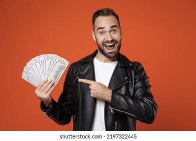 Excited young bearded man in white t-shirt, black leather jacket standing pointing index finger on fan of cash money in dollar banknotes looking camera isolated on orange background studio portrait