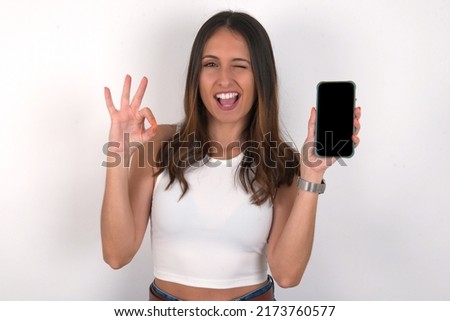Excited young beautiful caucasian woman wearing white top over white background showing smartphone blank screen, blinking eye and doing ok sign with hand.  Advertisement concept.